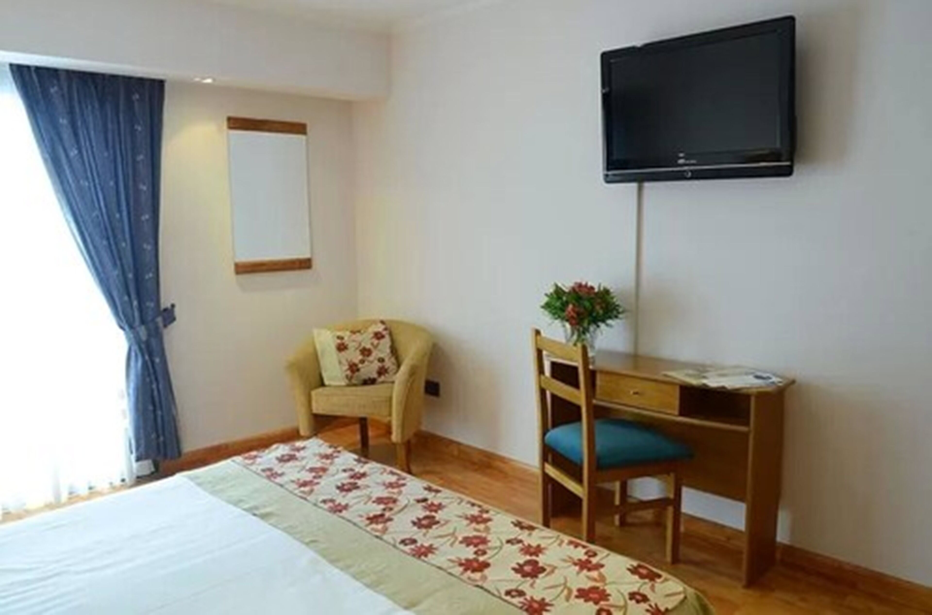 A room with a bed, a desk, a chair, a television mounted on the wall, and a window with curtains.
