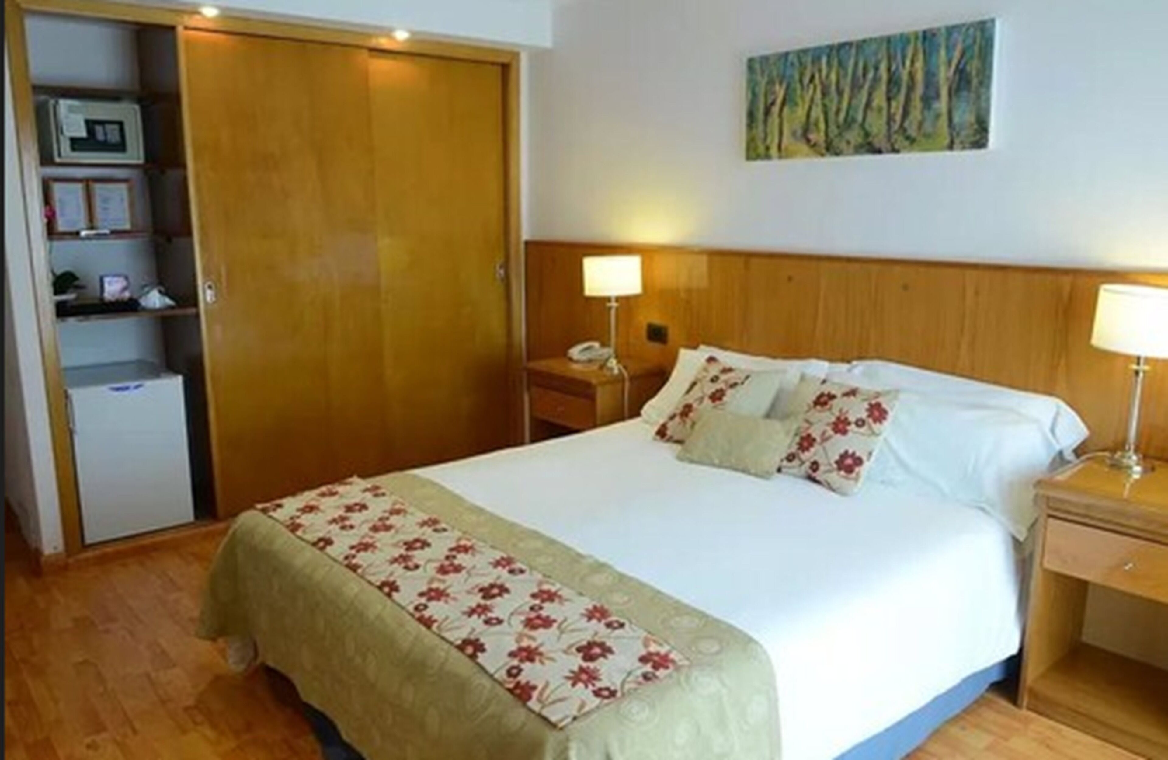 A hotel room with a bed, bedside tables, a wardrobe, and a painting on the wall.