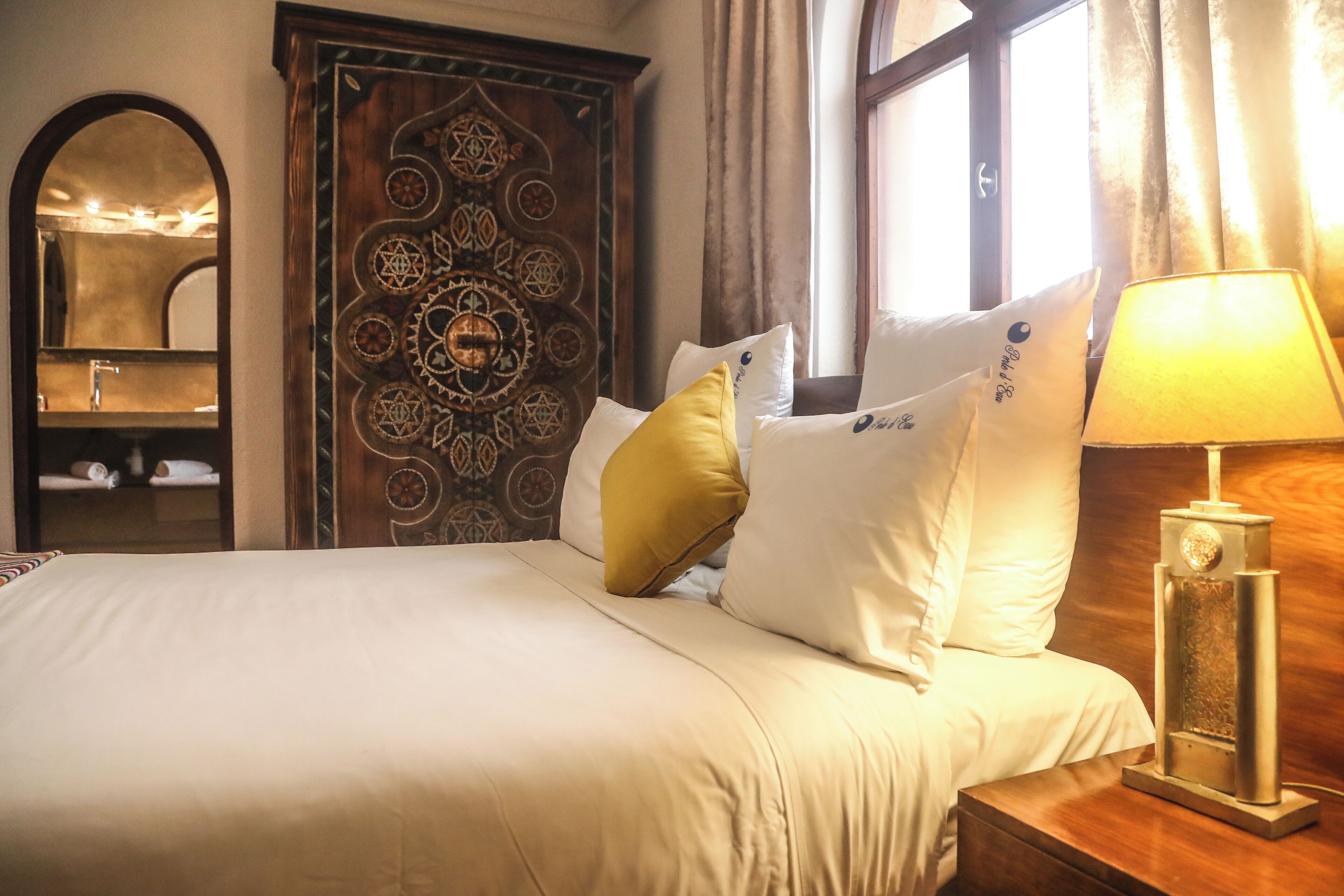 A neatly made bed with white linens and a gold accent pillow, a wooden nightstand with a lamp, and a decorative wooden door with intricate patterns.