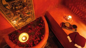 Un baño con una bañera llena de pétalos de rosa y velas, una bandeja con una taza y unos lentes de sol, y una toalla roja sobre un banco.