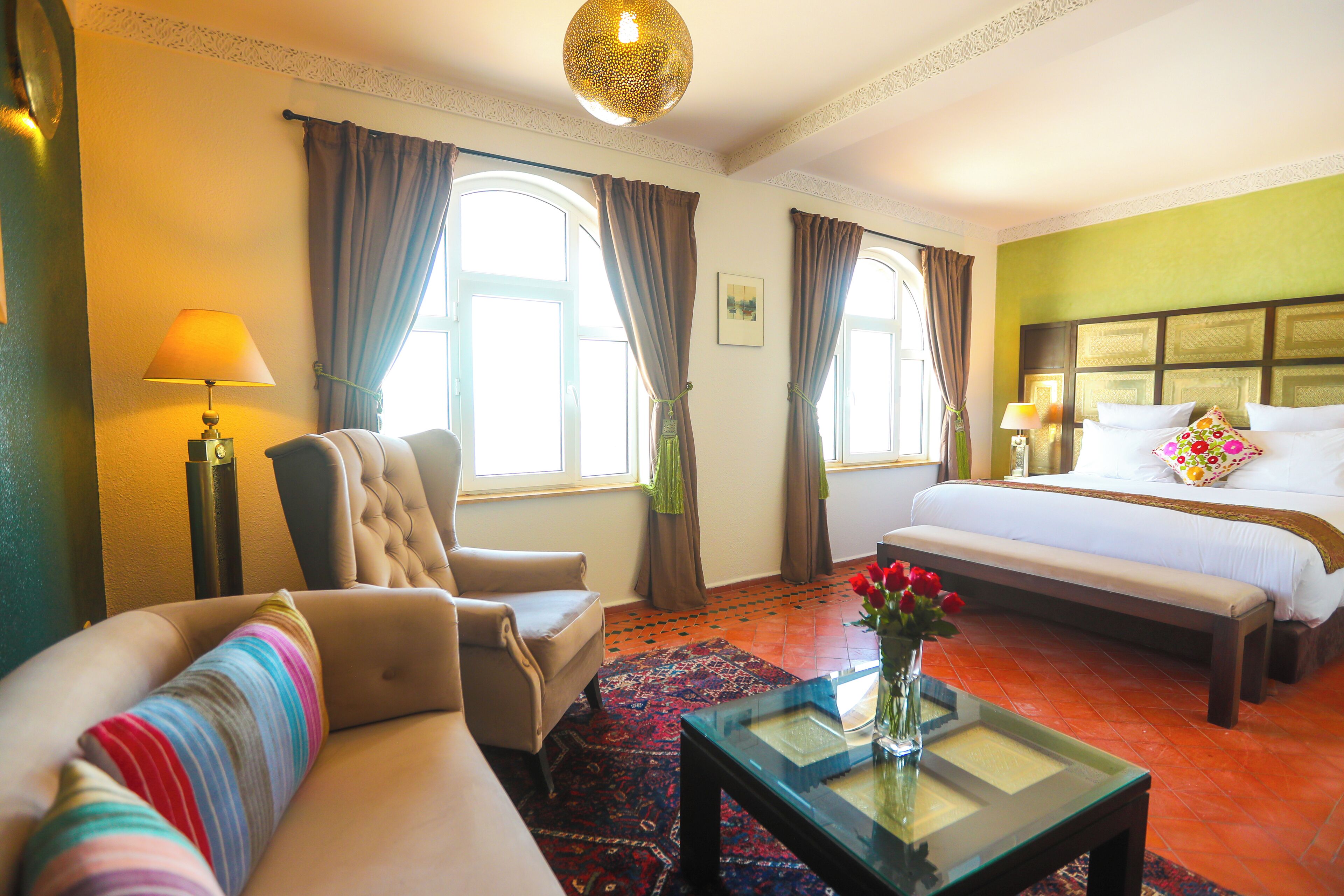A hotel room with a bed, a sofa, a chair, a table with a vase of flowers, and a window with curtains.