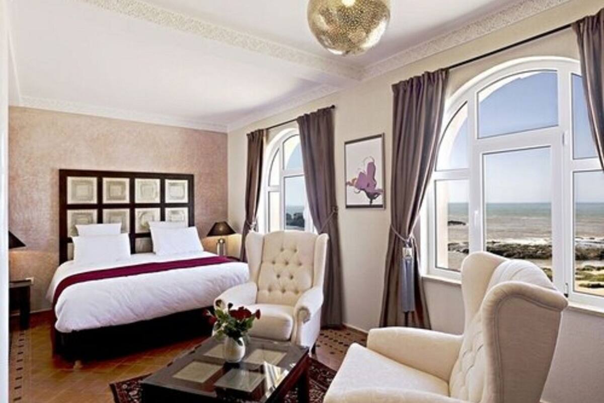 A hotel room with a large bed, a sitting area with a chair, a coffee table, and a view of the sea through large windows.