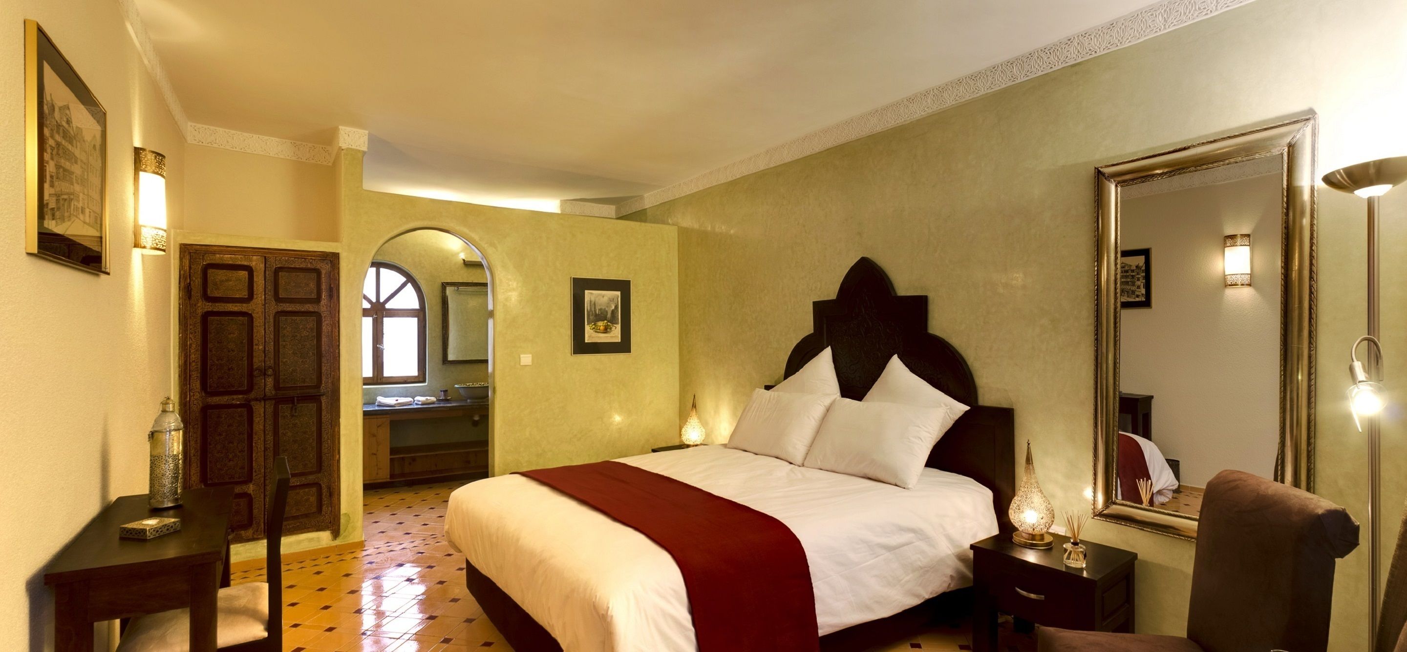 Una habitación de hotel con una cama grande, una puerta de madera, un espejo y una mesita con una lámpara.
