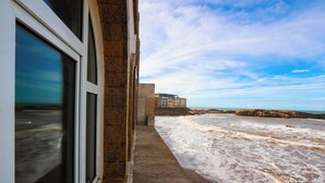 Vista costera desde una ventana con vista a una playa arenosa, olas, cielo despejado y un edificio a lo lejos.