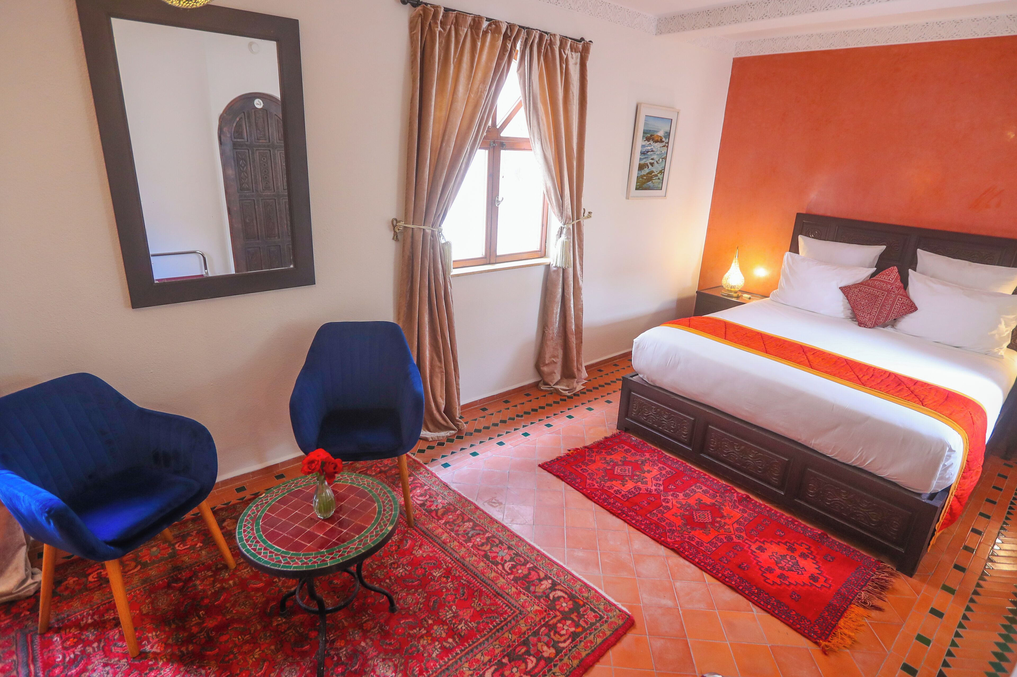 A hotel room with a bed, two blue chairs, a small table with a vase, a red rug, and a window with curtains.