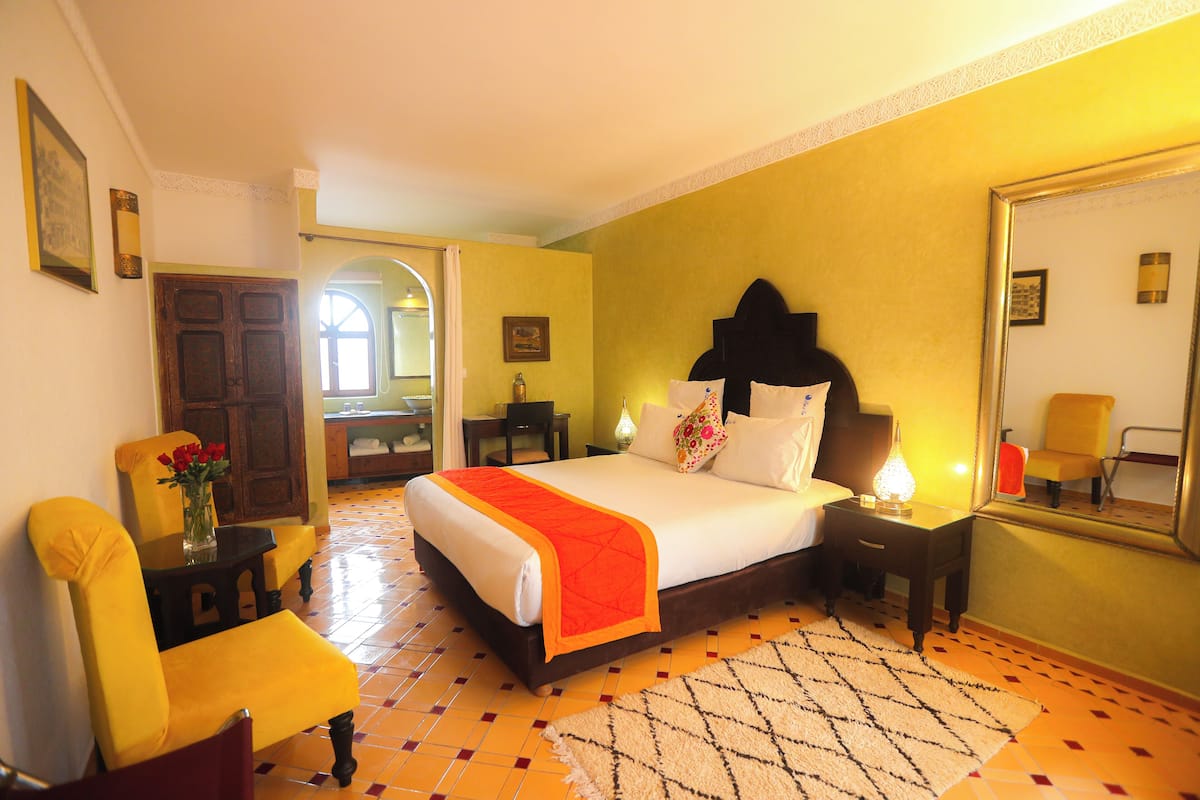 A hotel room with a large bed, a yellow armchair, a small table with a vase of flowers, and a mirror on the wall.