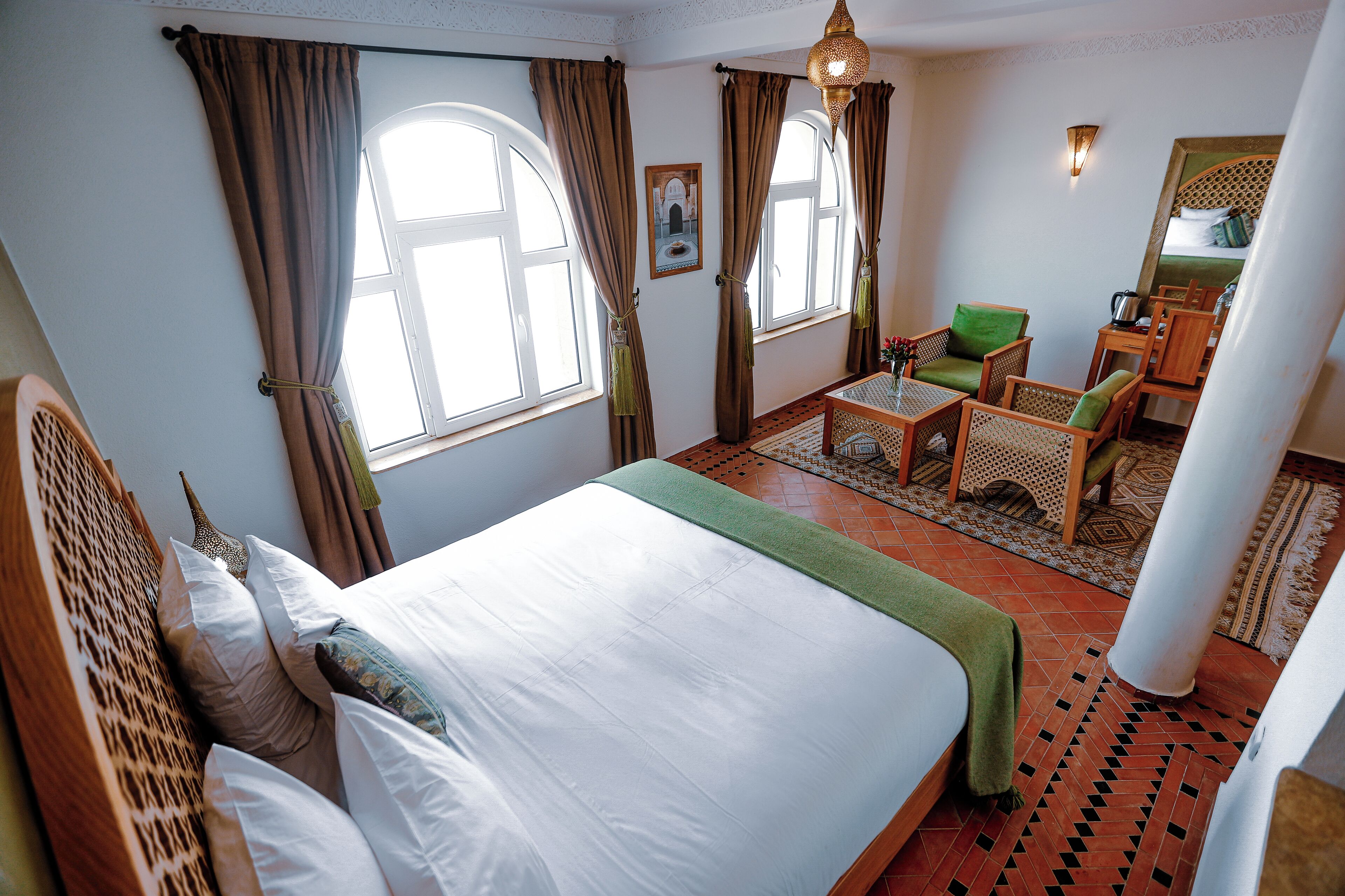 A hotel room with a large bed, a green armchair, a wooden coffee table, and a view of the outdoors through the windows.