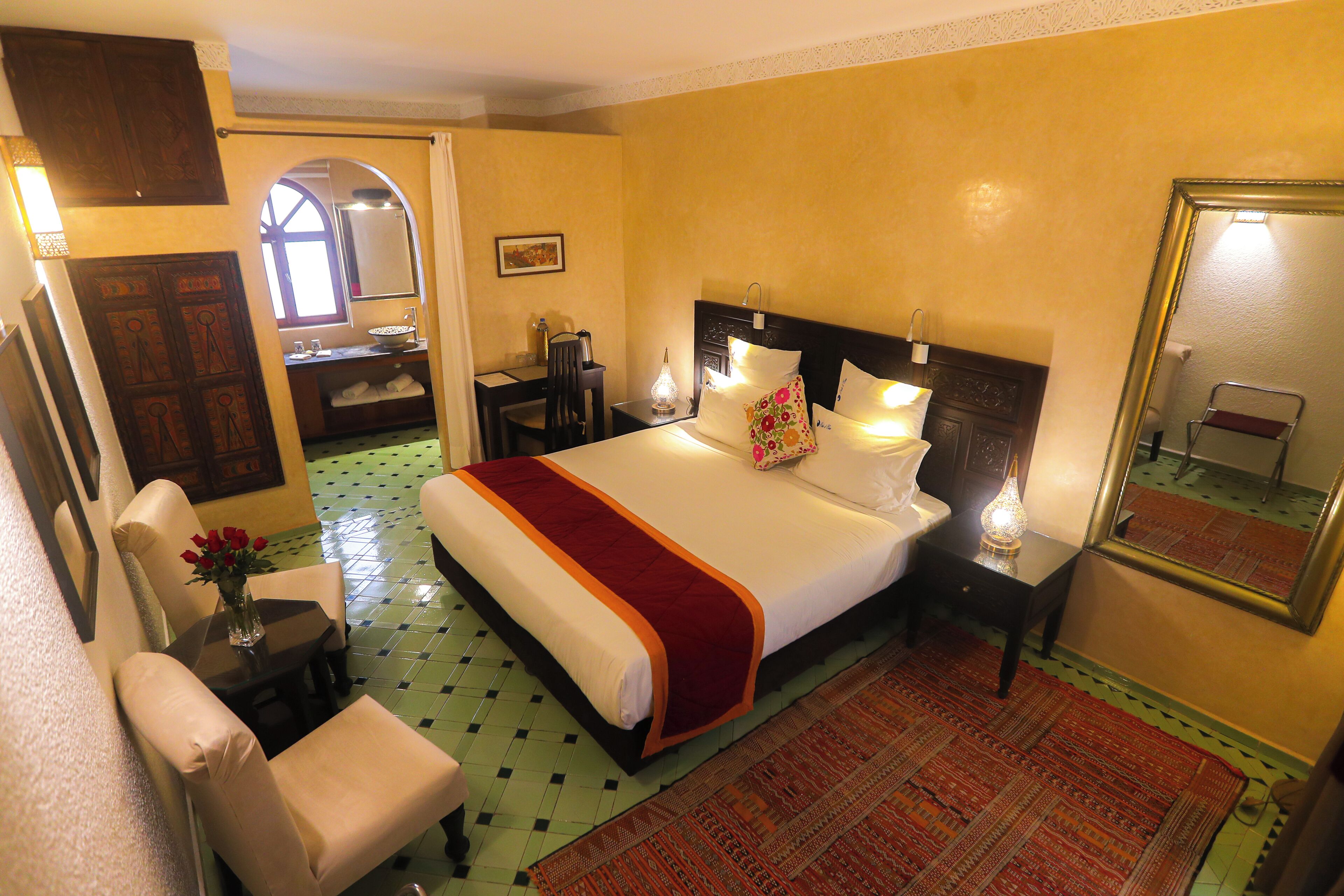 A hotel room with a large bed, a desk with a chair, a small table with a vase of flowers, and a bathroom visible through an archway.