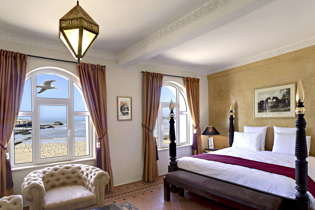 A bedroom with a large bed, two windows with curtains, a chair, a lamp, and a clock.