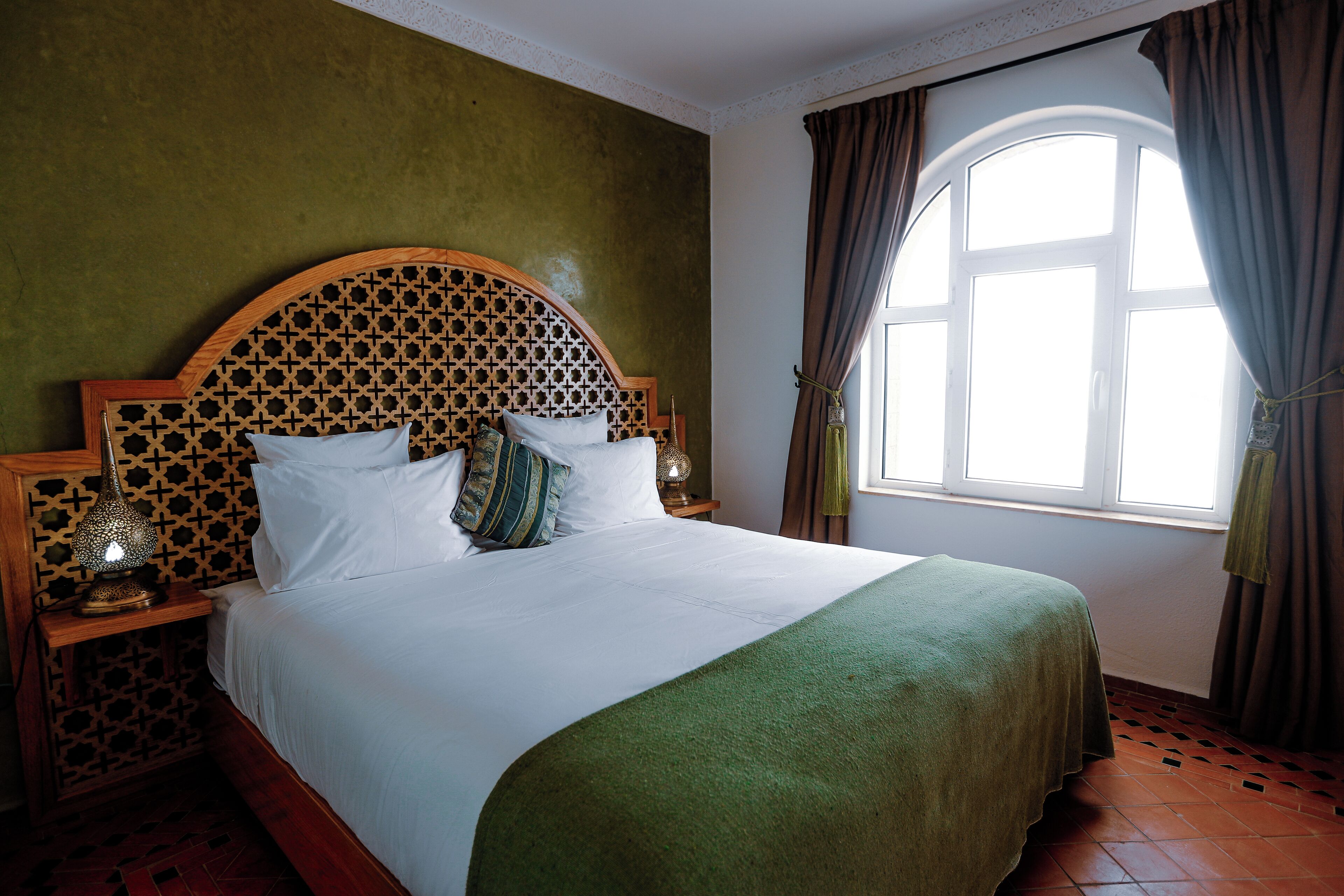 A neatly made bed with a green blanket, a wooden headboard with a geometric pattern, and a nightstand with a lamp.