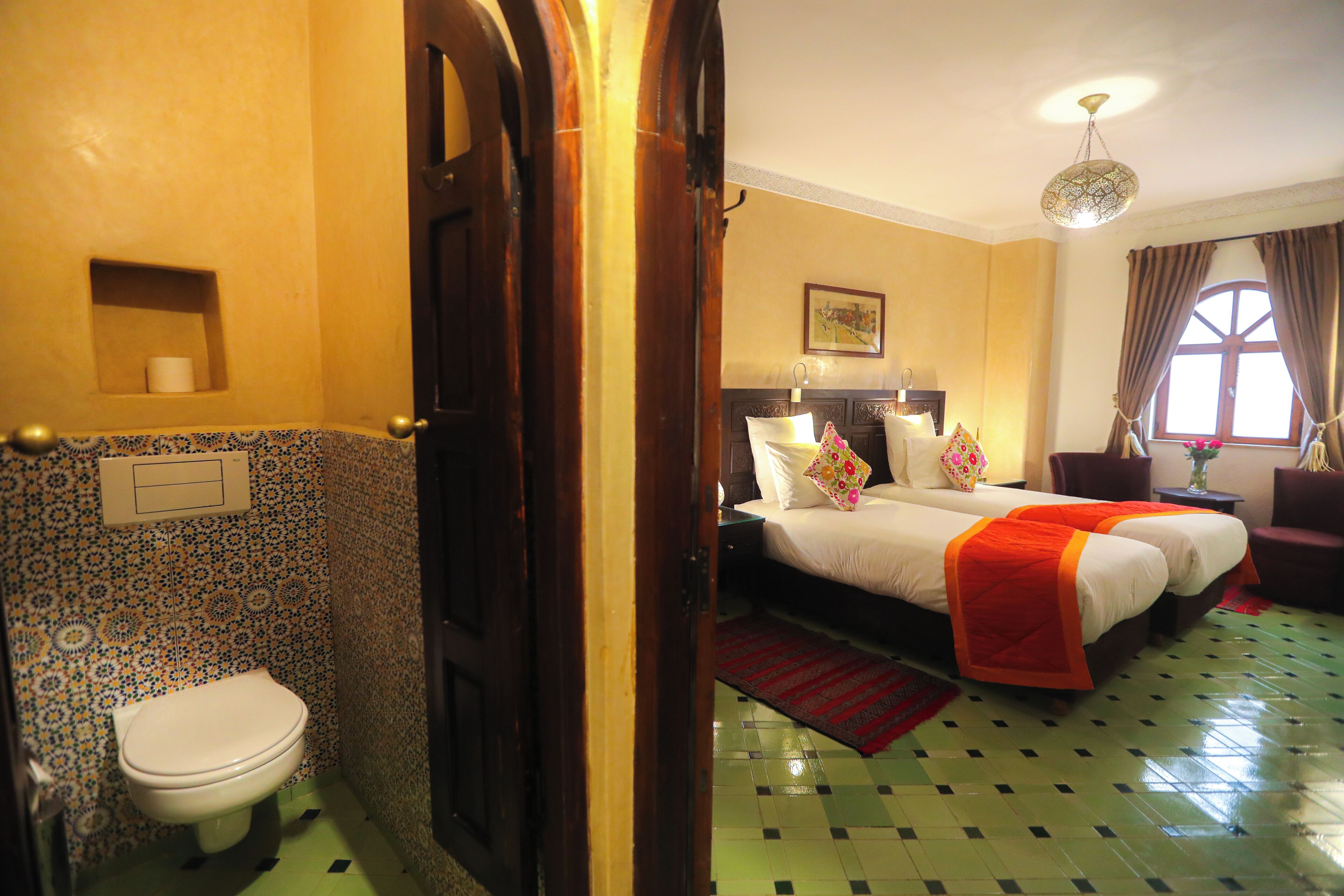 Ein Hotelzimmer mit Toilette, einem Bett mit roter Decke, einem Chandelier und einem Fenster mit Vorhängen.