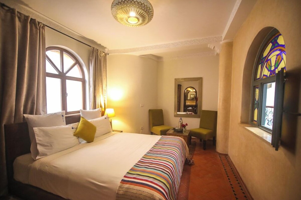 A hotel room with a bed, a chair, a small table, a mirror, and a window with curtains.