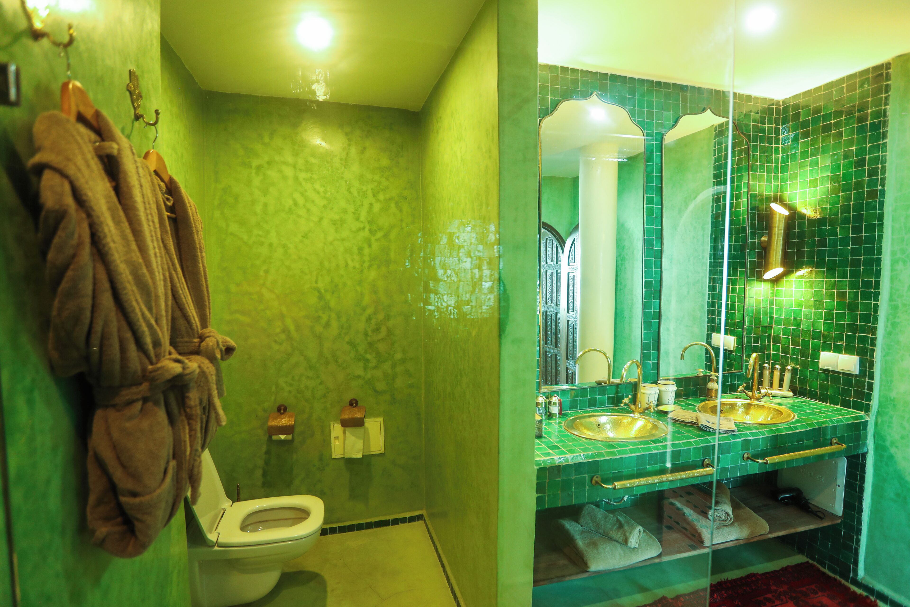 A bathroom with a green tiled wall, a white toilet, a glass-enclosed sink area, and a hanging bathrobe.