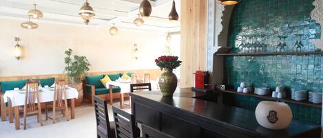 Interior de restaurante con mesas y sillas de madera, lámparas colgantes, un jarrón decorativo y un reverso de azulejos.