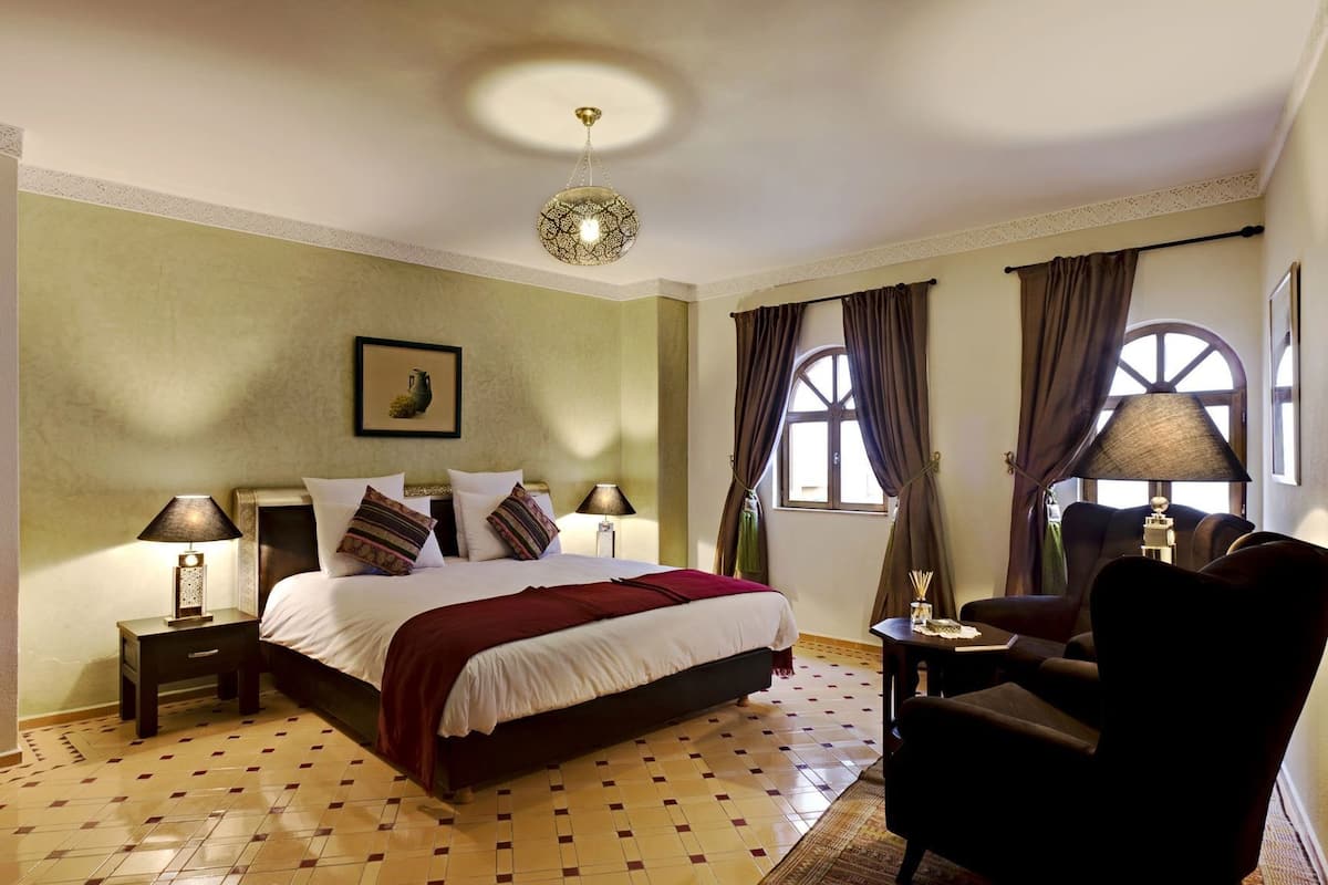 A hotel room with a bed, two bedside tables with lamps, a chair, a window with curtains, and a ceiling light fixture.