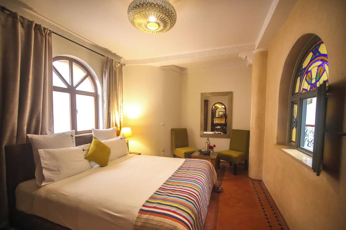 A hotel room with a large bed, a chair, a small table, and a window with a stained glass design.