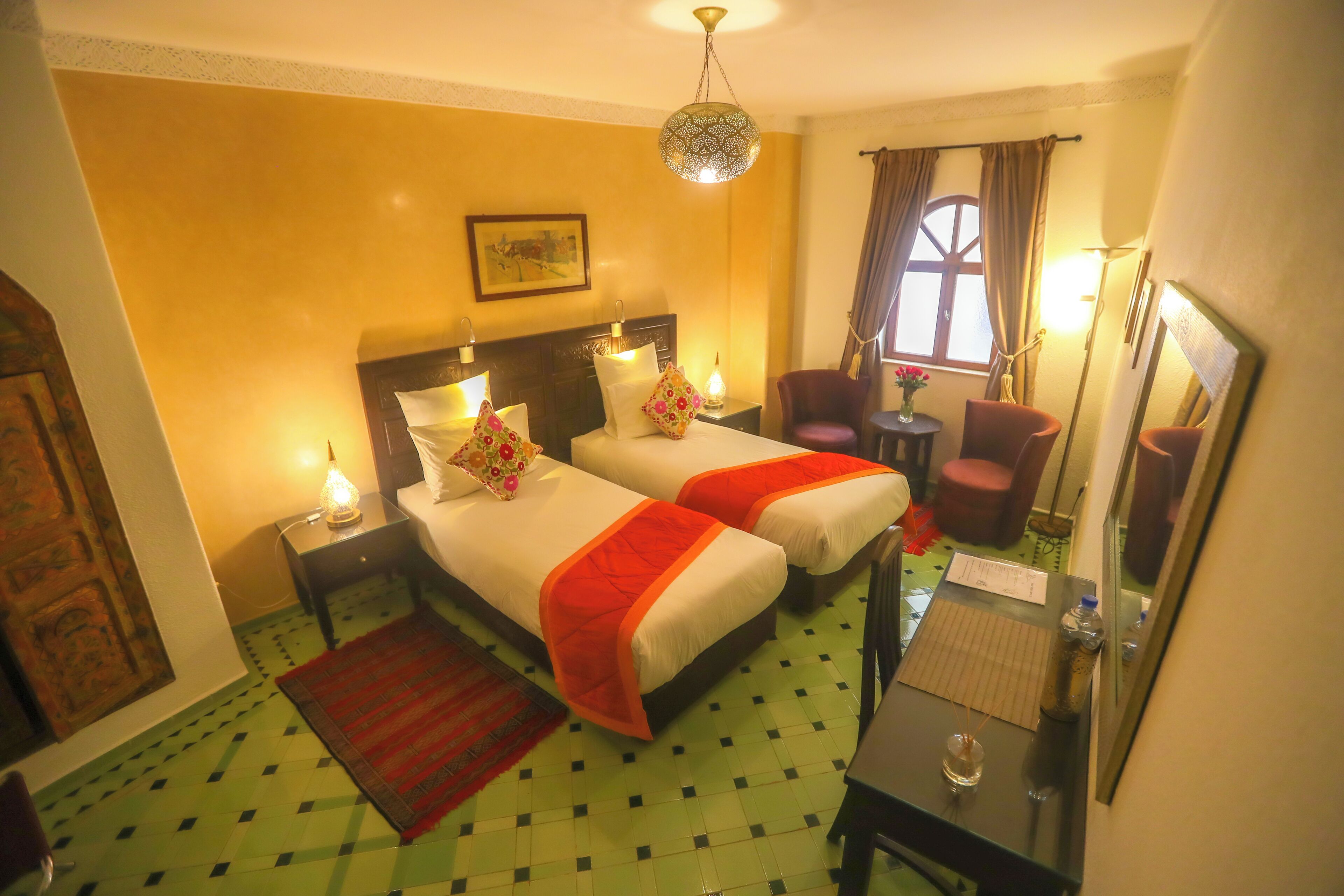 A hotel room with two beds, a sitting area with a chair and table, a mirror, and a window with curtains.