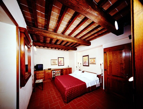 A room with a wooden ceiling, a bed with a red bedspread, a desk with a chair, and a wooden door. - Fattoria Pianetti (Manciano)