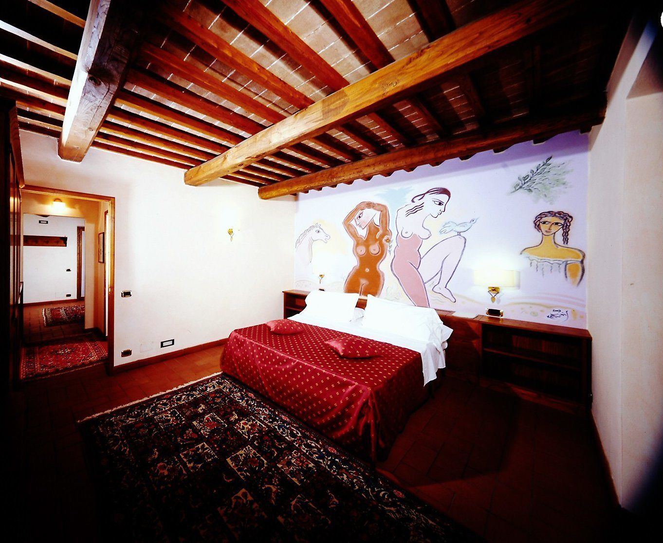Une chambre avec un plafond en bois, un lit recouvert d’une courtepointe rouge, un tapis à motifs et un mur orné d’une fresque.