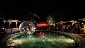 Piscina exterior iluminada de noche con una gran esfera transparente en el centro, rodeada de gente y sombrillas.