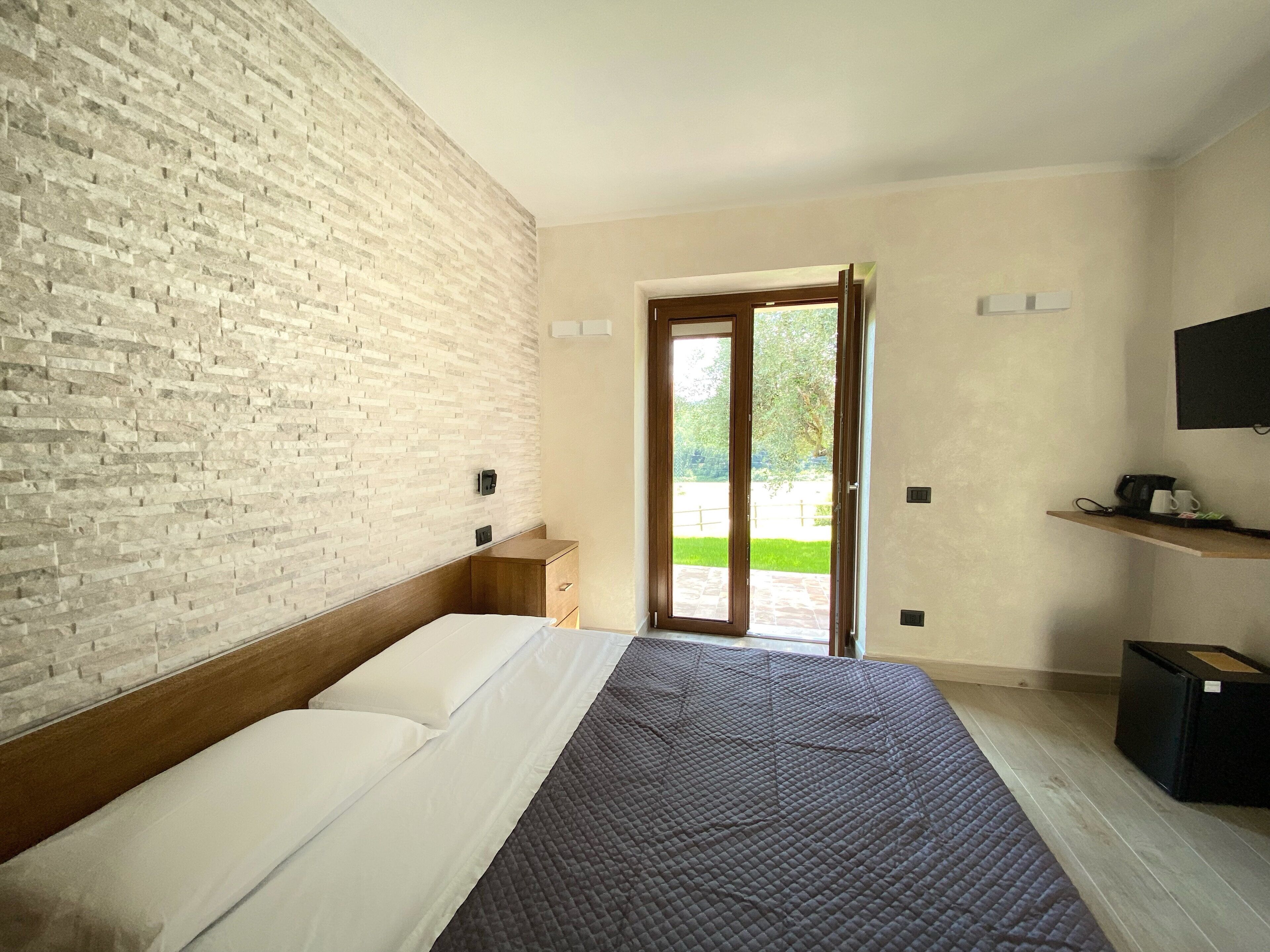 Een slaapkamer met een bakstenen muur, een bed met witte lakens, een houten nachtkastje, een schuifdeur naar een tuin en een tv aan de muur.