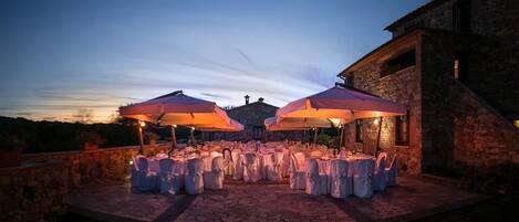 An outdoor dining area with round tables and chairs, covered by large umbrellas, set against a stone building with a view of the hills.
