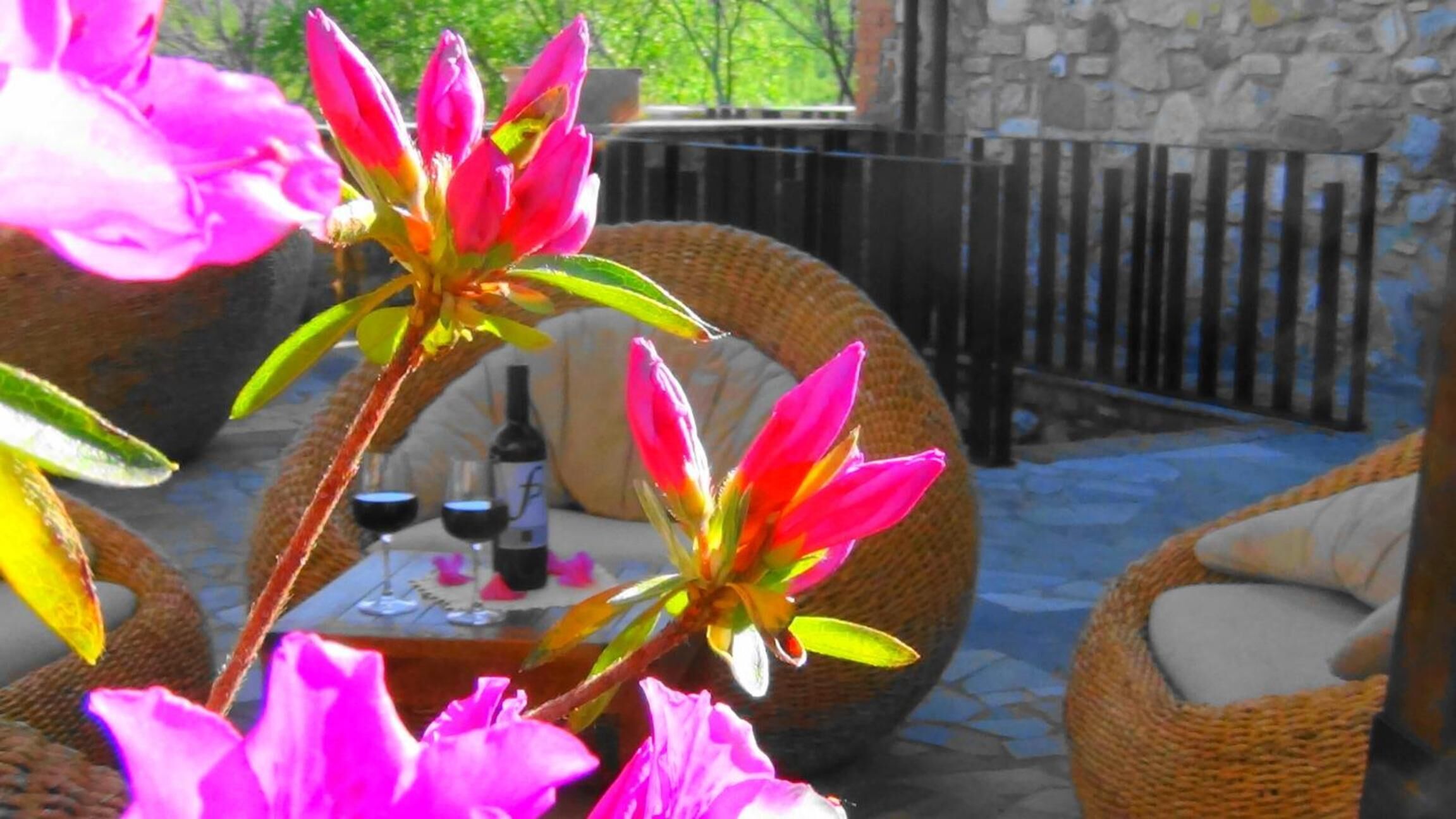Wicker furniture with cushions, a table with a bottle and glasses, and a stone wall in the background.