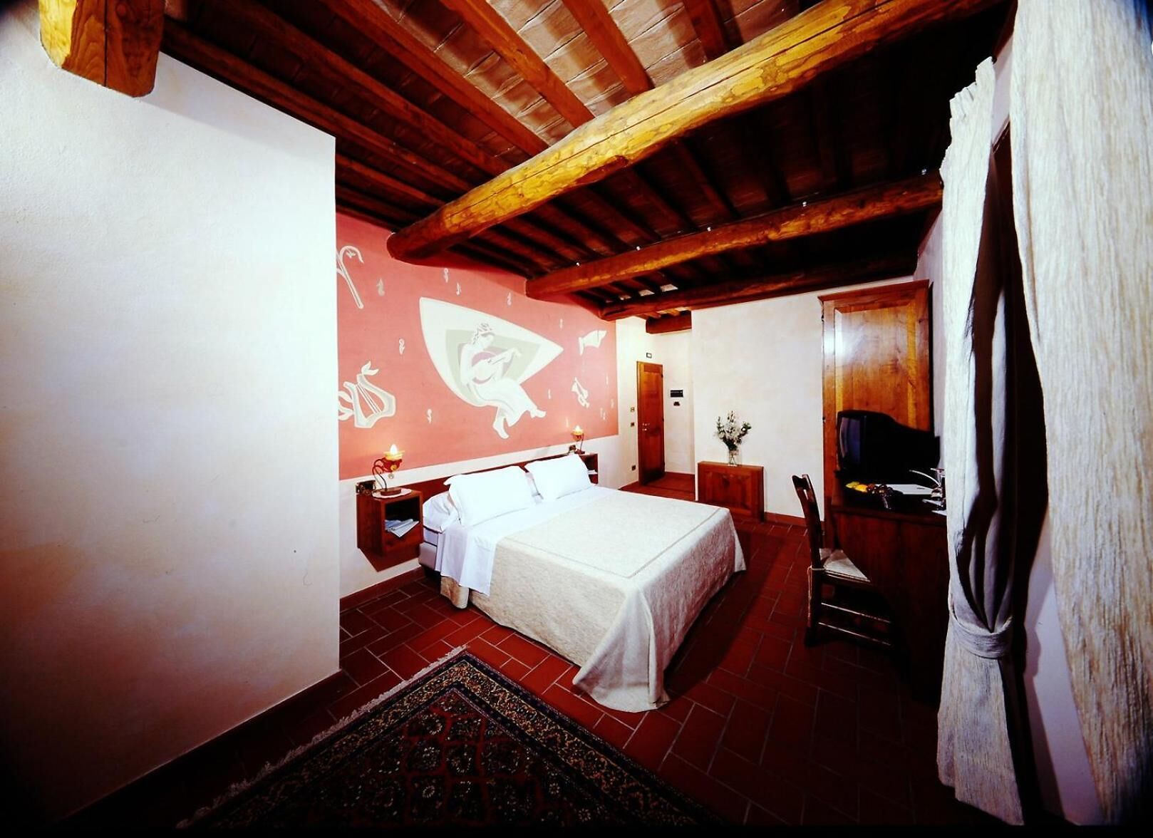 A bedroom with a large bed, wooden ceiling beams, a red wall with white patterns, a small bedside table, and a chair.