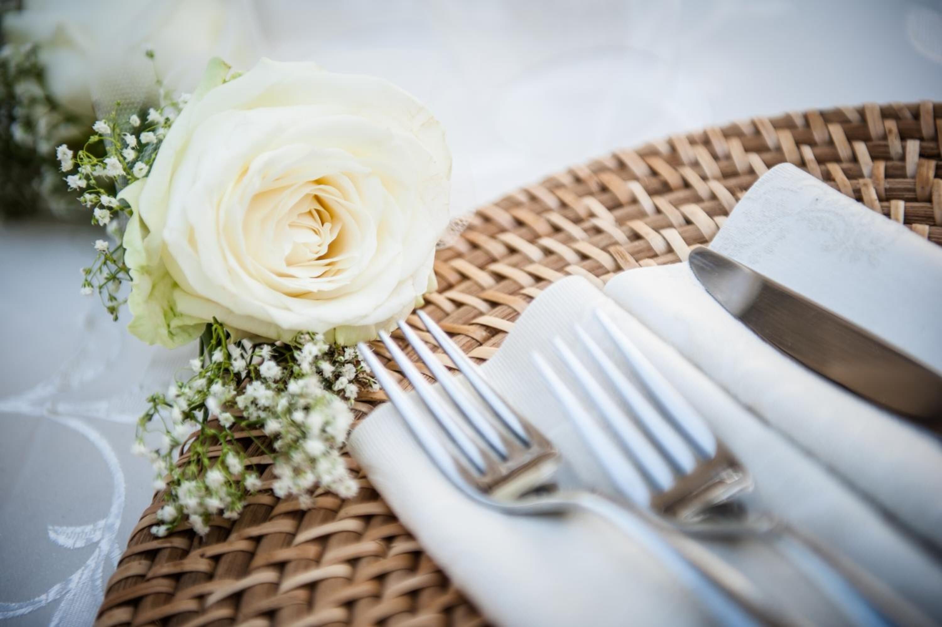 A round wicker table with a white rose and small white flowers, accompanied by a folded napkin and a fork.