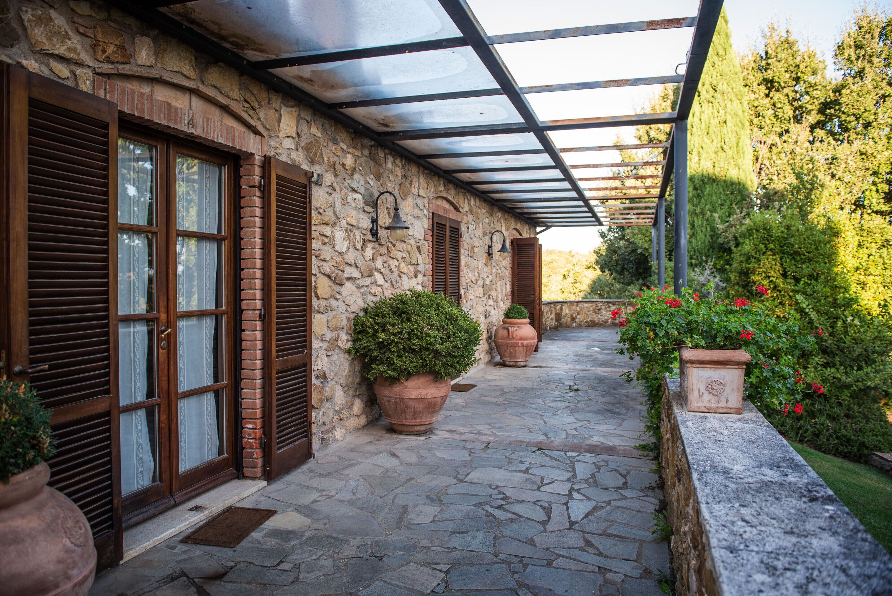 Un edificio de piedra con un porche cubierto, ventanas grandes con contraventanas y plantas en macetas.