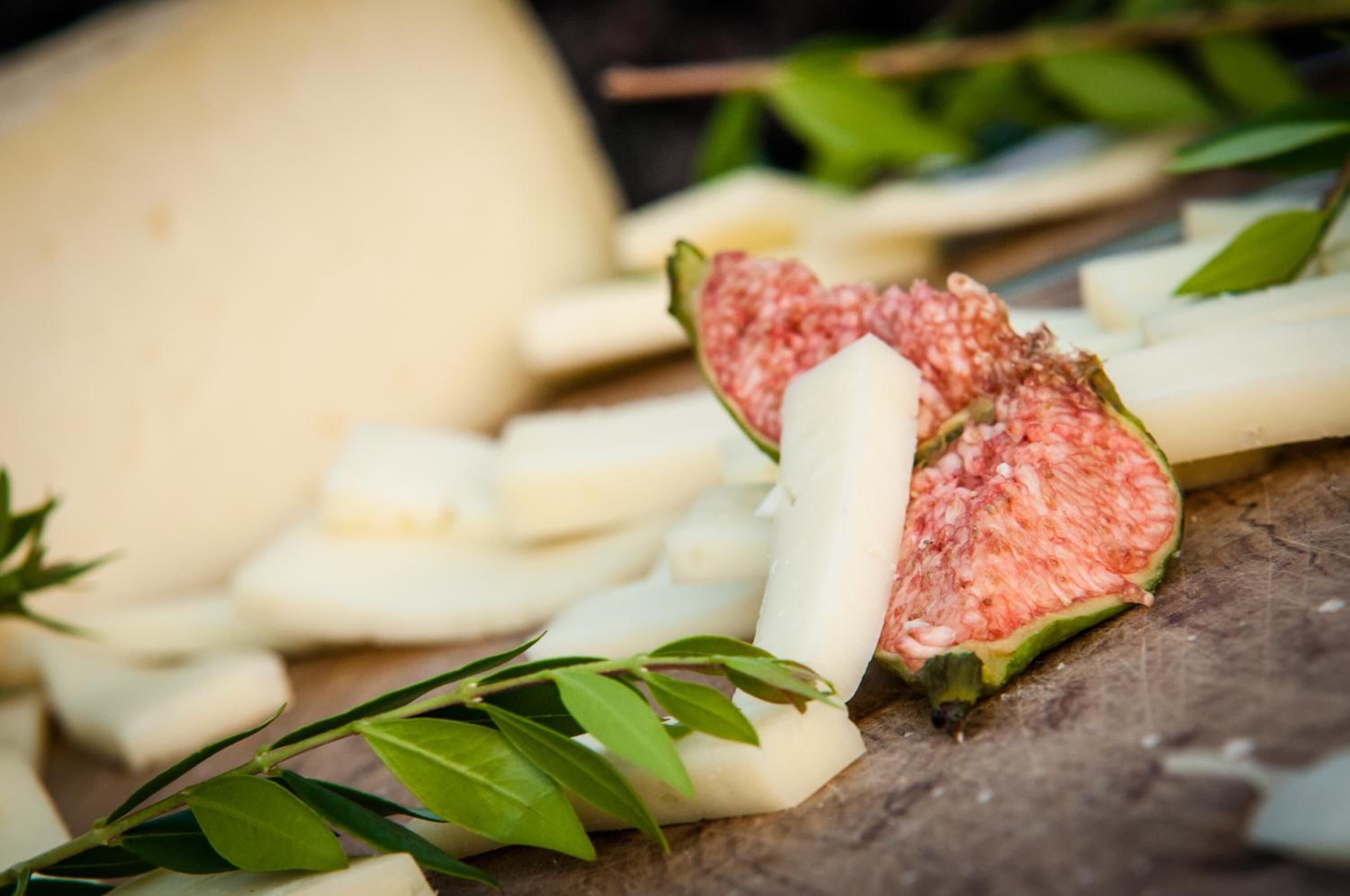 A close-up of various food items, including a whole fig, sliced figs, and a white cylindrical object, possibly cheese, on a wooden surface with green leaves.