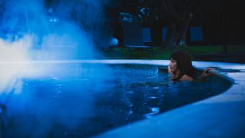 A person in a hot tub with steam rising from the water, surrounded by trees and a bench in the background.