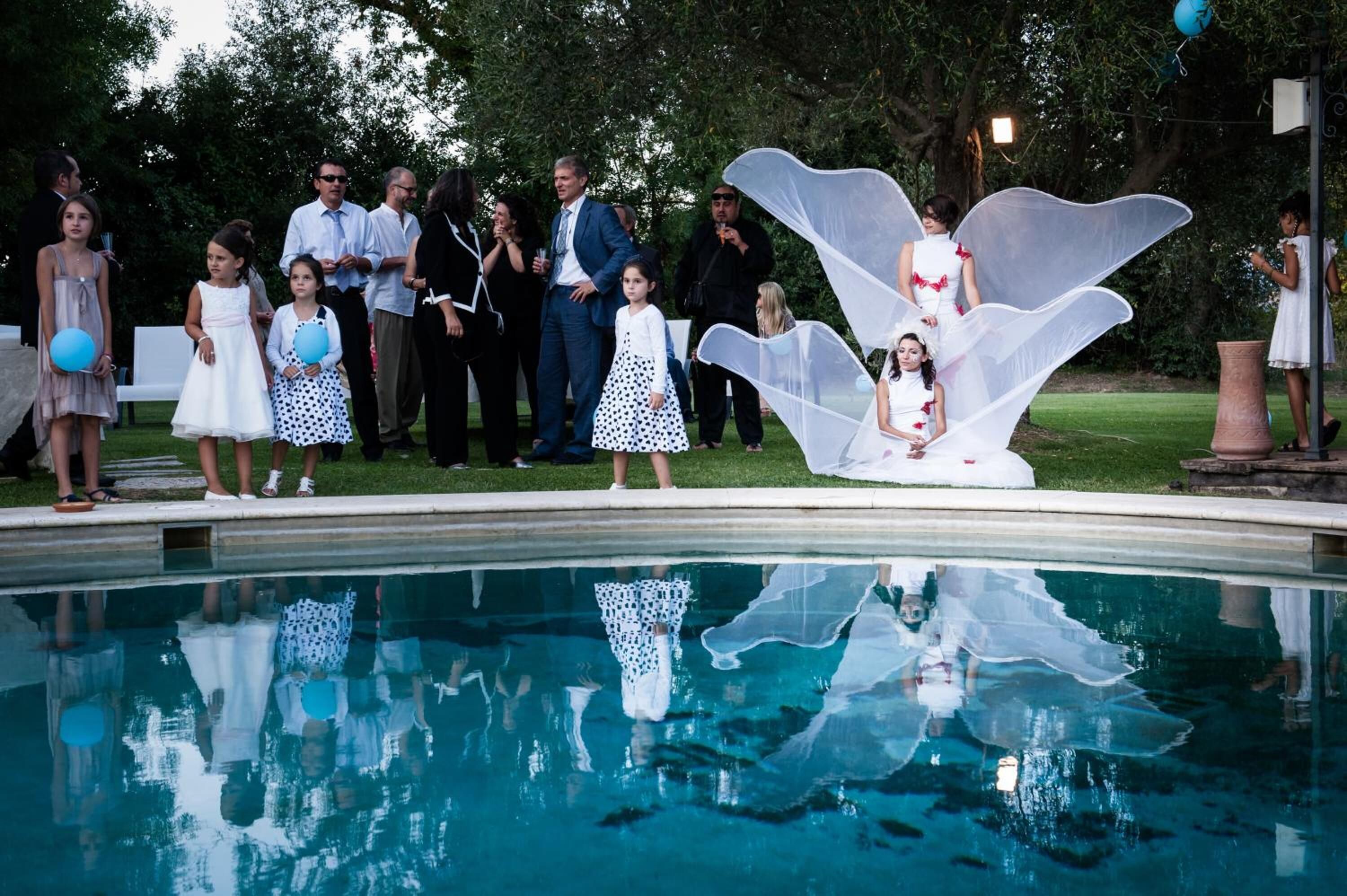 A poolside gathering with people standing by the pool, some holding balloons.