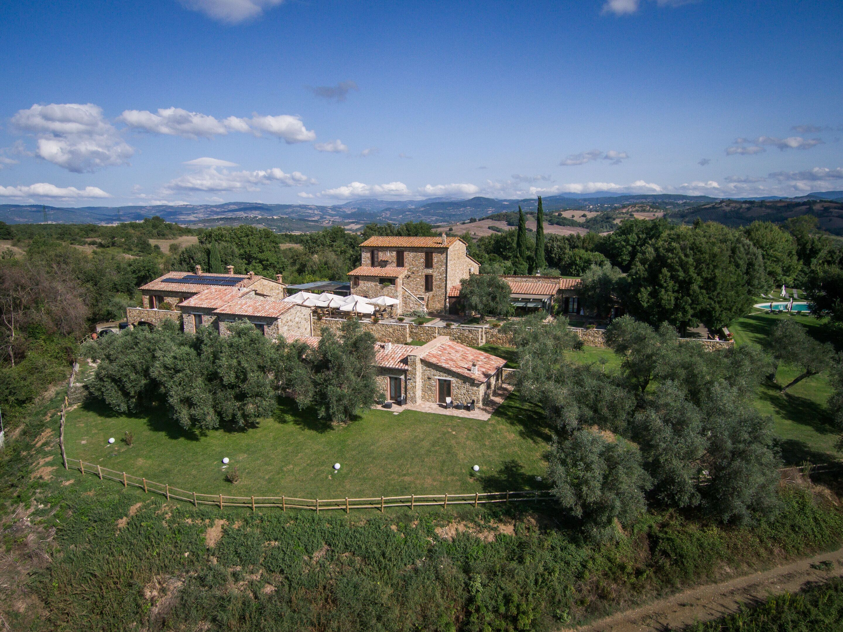 A stone villa with multiple buildings, surrounded by lush greenery and a fenced lawn.