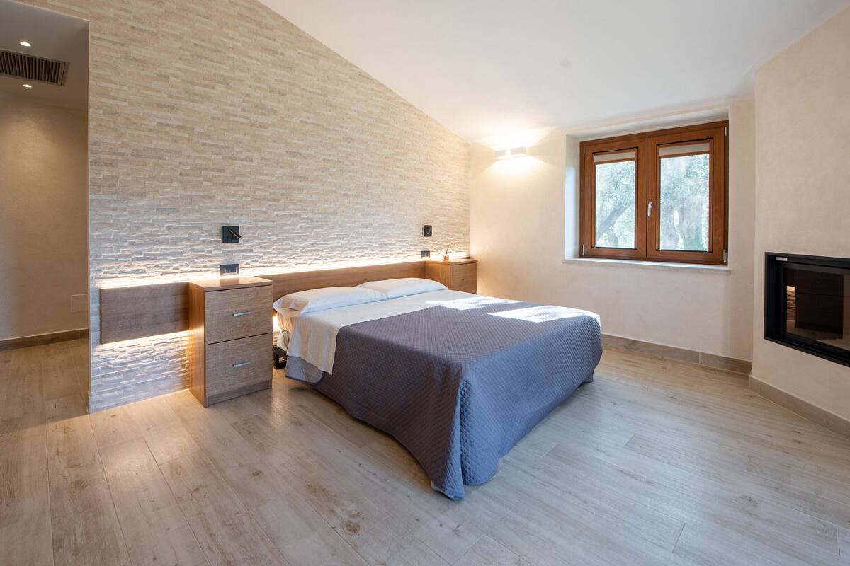 Un dormitorio con cama, mesita de noche, cómoda, pared de ladrillo, ventana y chimenea.