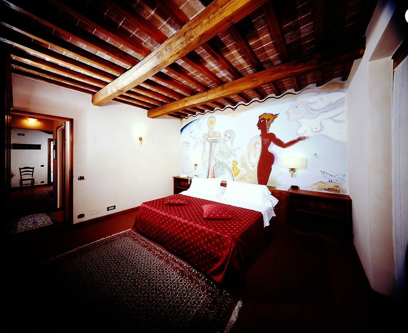 A bedroom with a wooden ceiling, a bed with red bedding, a nightstand, and a wall mural.
