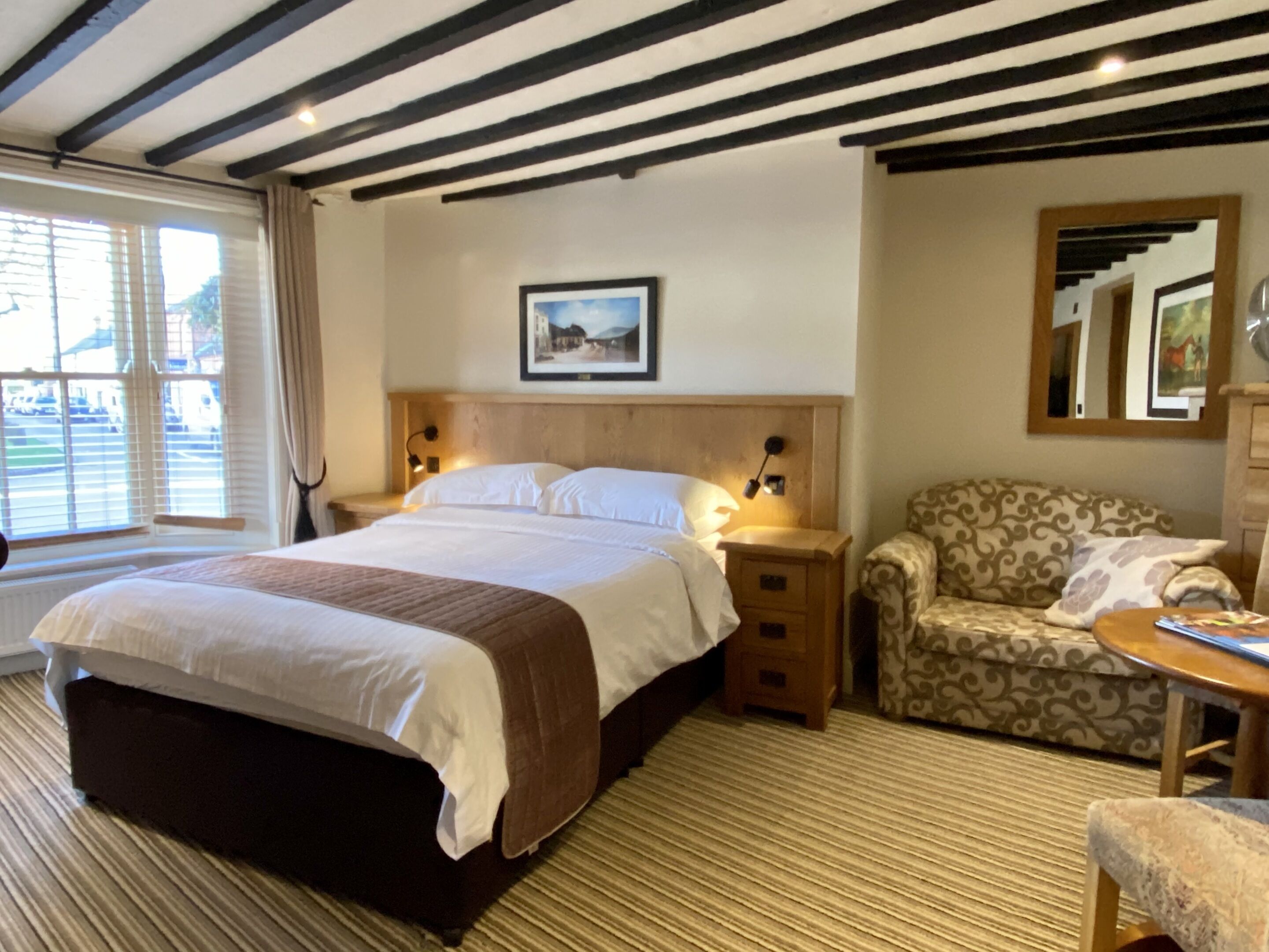 Una habitación de hotel con una cama grande, una mesita de noche de madera, un sillón estampado y una pequeña mesa con una lámpara.