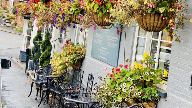 A pub with outdoor seating, hanging flower baskets, and a signboard listing room names and contact information.