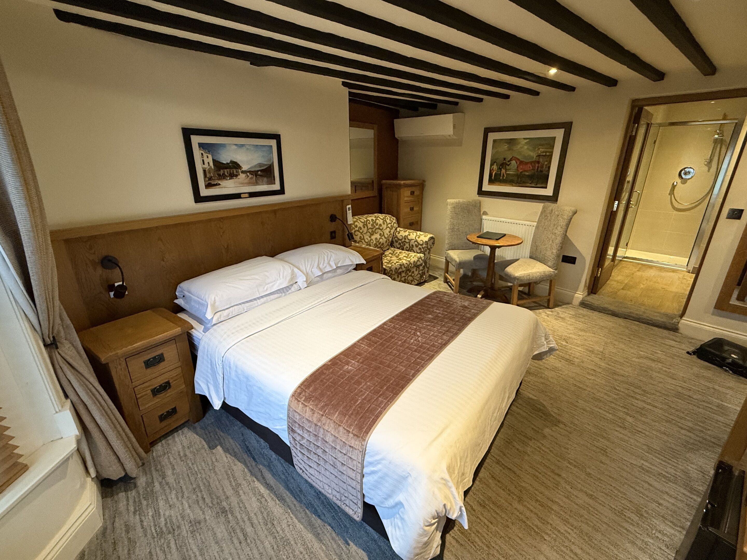 Habitación de hotel con una cama grande, cabecera de madera, dos cuadros en la pared, una mesita con una silla y una zona de ducha.