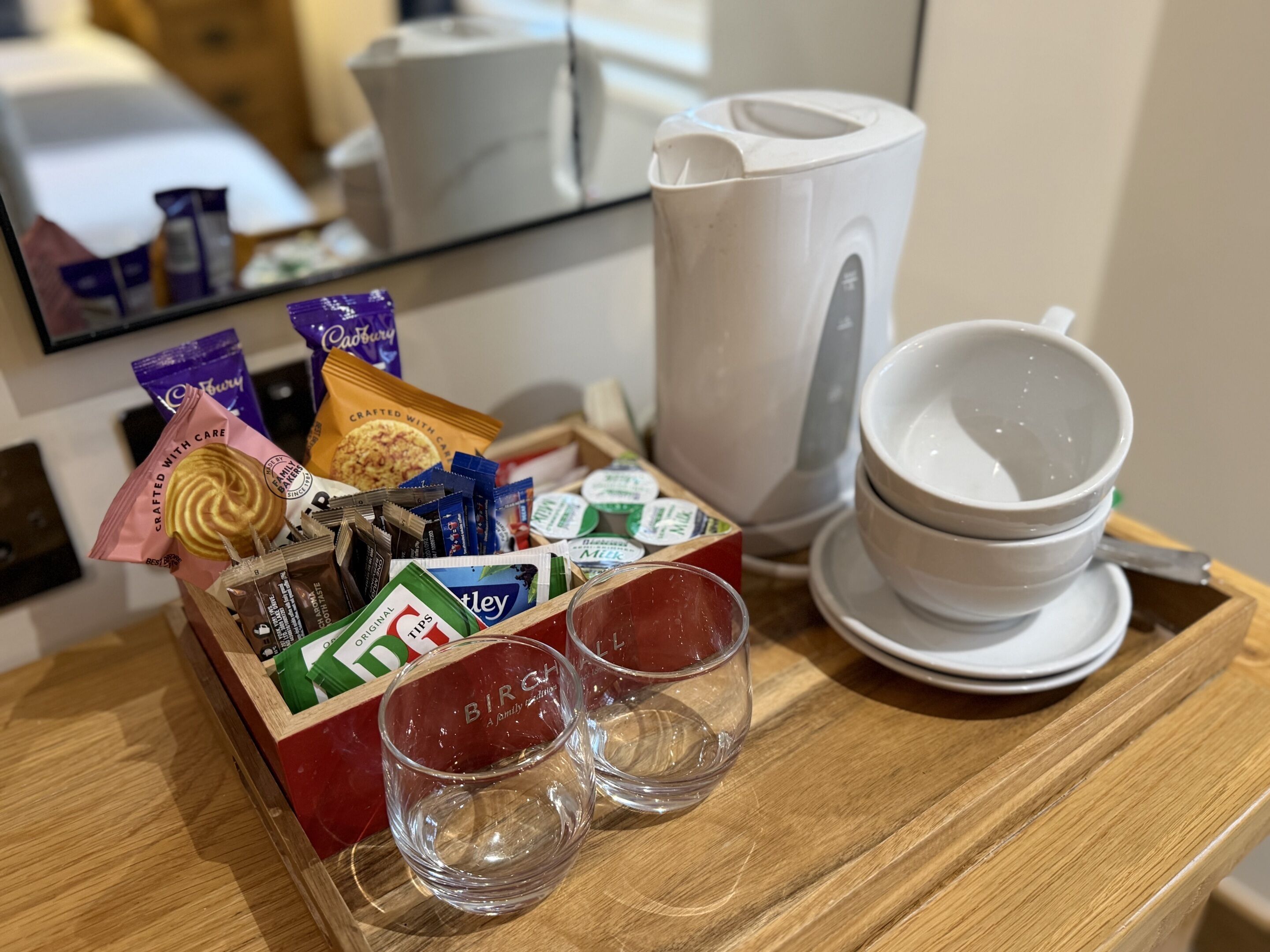 Een dienblad met diverse snacks, een witte koffiezetapparaat en twee glazen op een houten tafel.
