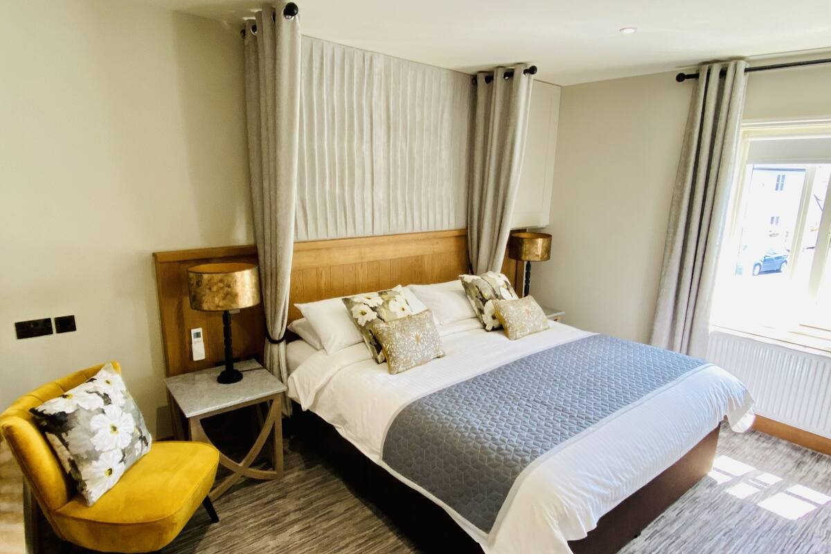 A hotel room with a large bed, a wooden headboard, a nightstand with a lamp, a yellow chair, and a window with curtains.