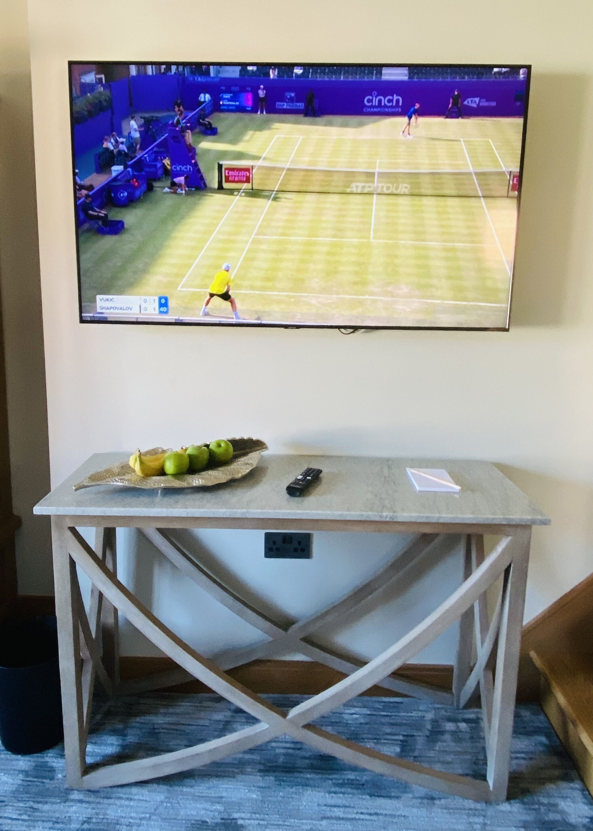 Un televisor montado en la pared muestra un partido de tenis. Debajo del televisor, hay una mesa con un plátano, un control remoto y un papel.