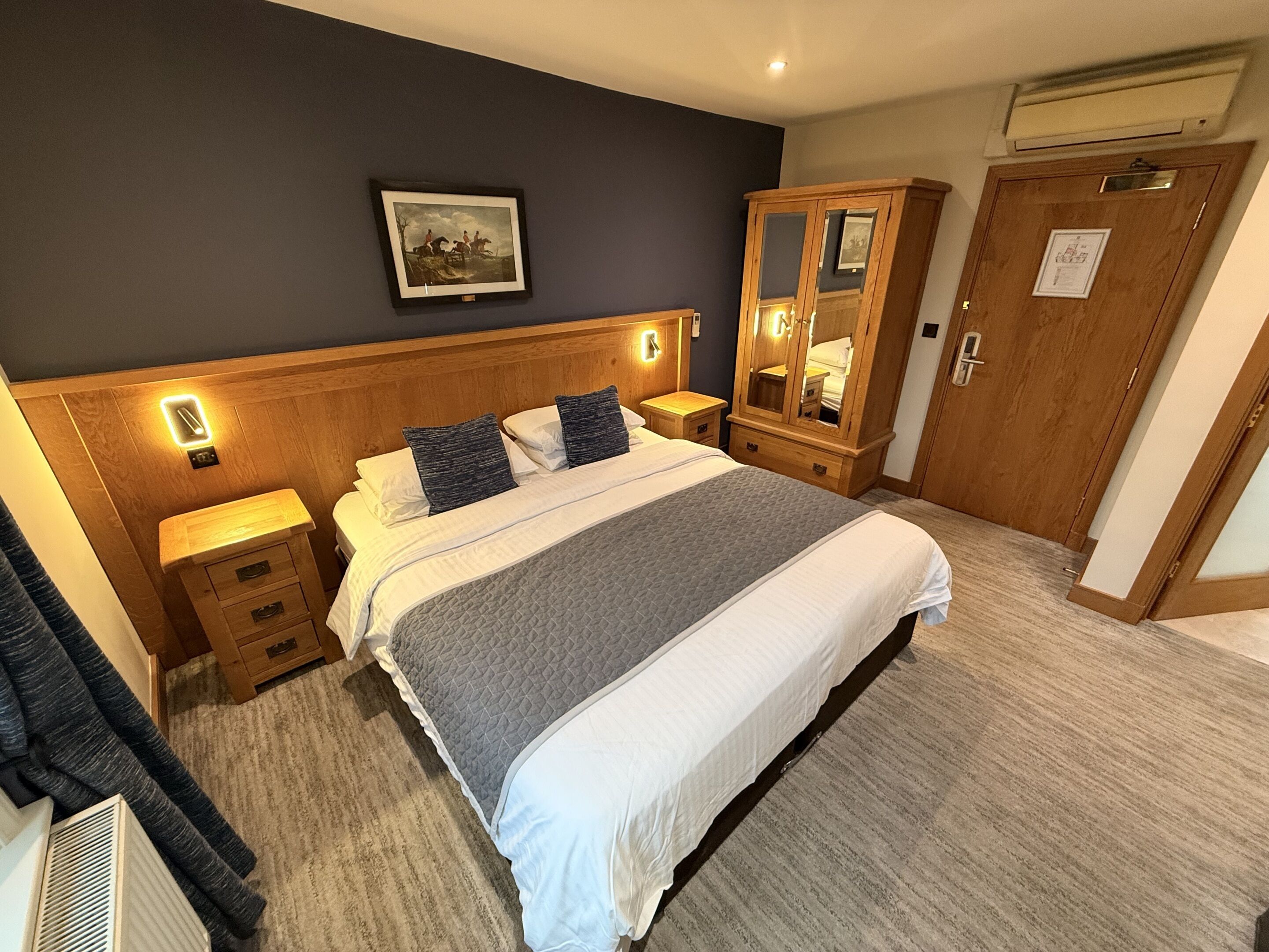 Quarto de hotel com uma cama grande, cabeceira de madeira, mesas de cabeceira com luminárias, guarda-roupa e um quadro emoldurado na parede.