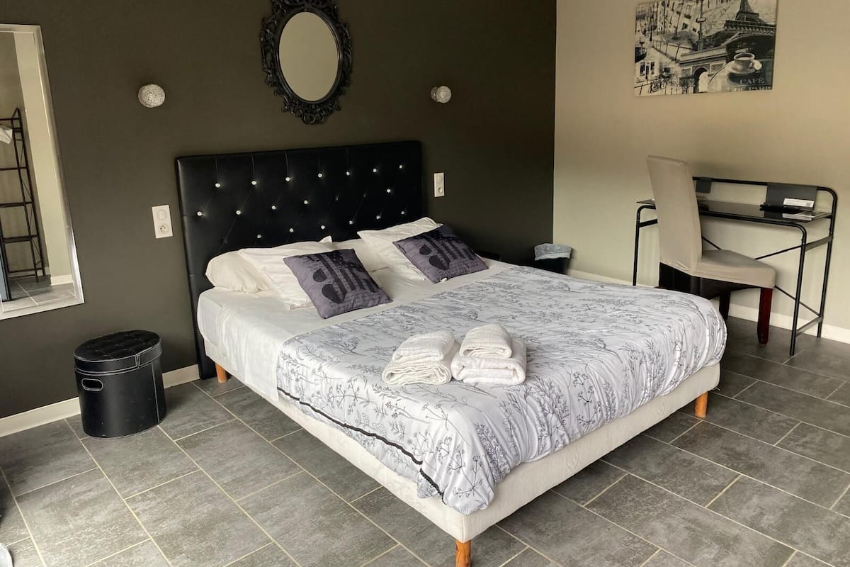 A bedroom with a bed, a desk, a chair, a trash bin, and a wall-mounted mirror.
