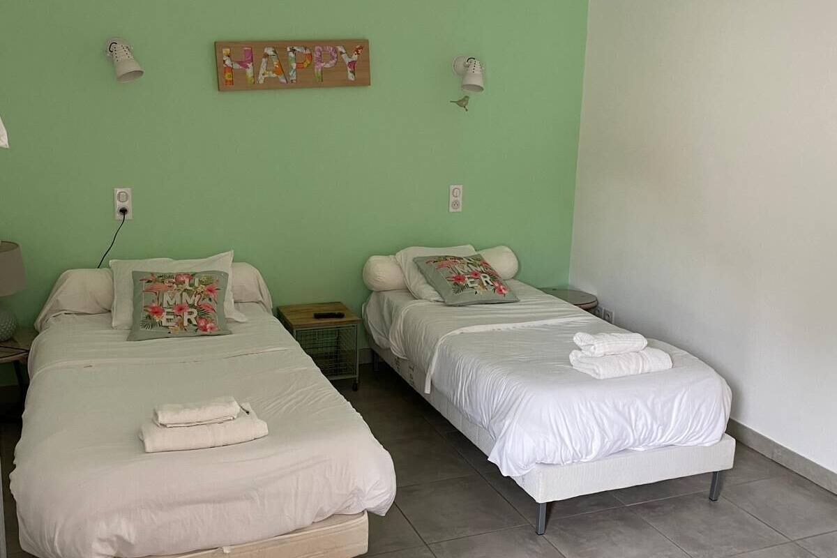A room with two single beds, a green wall, a wooden headboard, a small table, and a hanging light fixture.