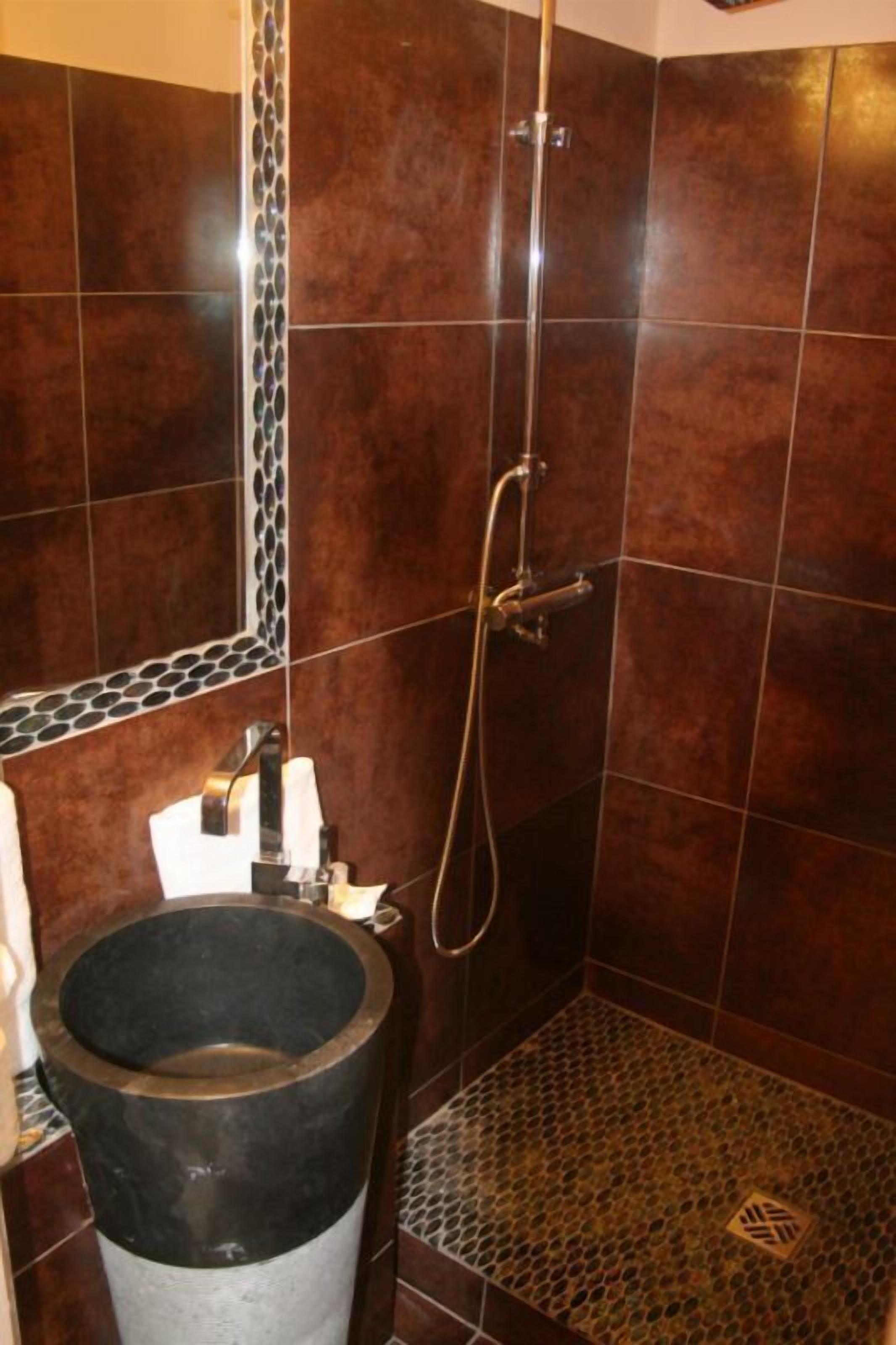Une salle de bain avec des carreaux de couleur brun foncé, un lavabo noir et une douche équipée d’un flexible.