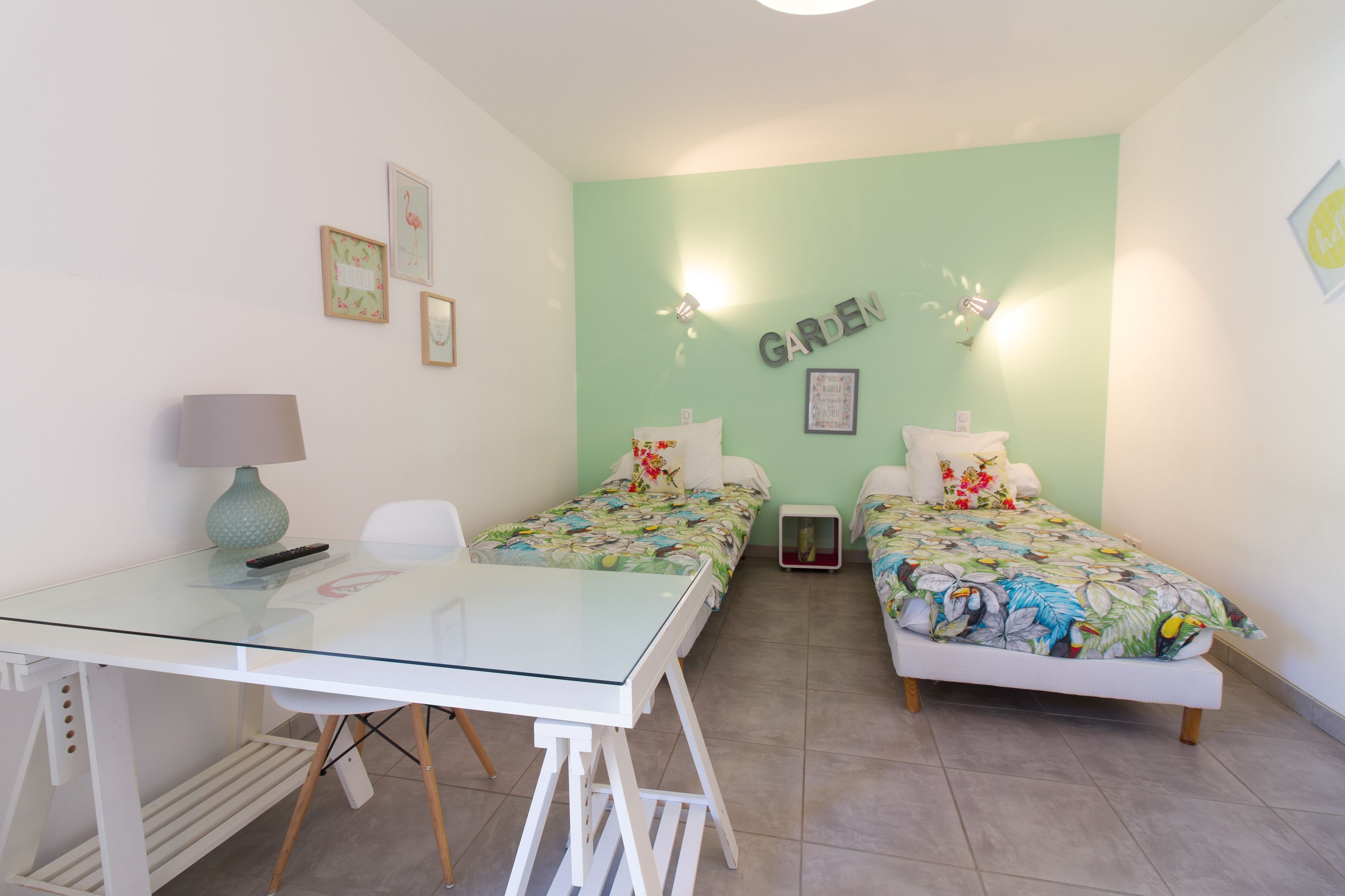 Ein Zimmer mit zwei Betten, einem Glastisch, einer Lampe und einer Wand mit dem Schriftzug 'GARDEN'.