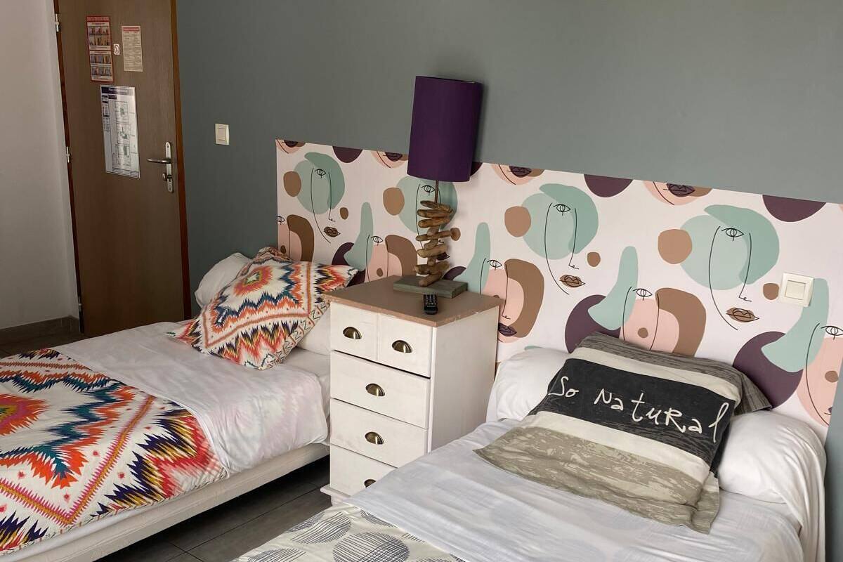 A bedroom with two beds, a nightstand, a wall-mounted lamp, and a door with a bulletin board.