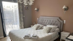 A neatly made bed with pillows and folded towels, a chandelier, and wall sconces in a room with a large window and a view of the outdoors. - Auberge Les Galets Hotel Restaurant (Peyruis)