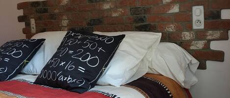 Una cama con cabecera de ladrillo, varios almohadones con ecuaciones matemáticas, y un cartel en la pared que dice "NEW YORK".